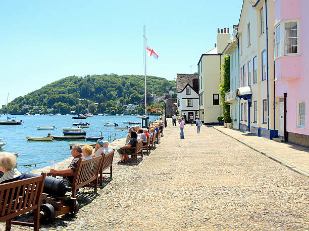 Dartmouth walkway with the sea on the left-hand side with people sat down appreciating the view and a flag sailing. On the right-hand side is a line of brightly painted houses (Yellow, blue and pink).