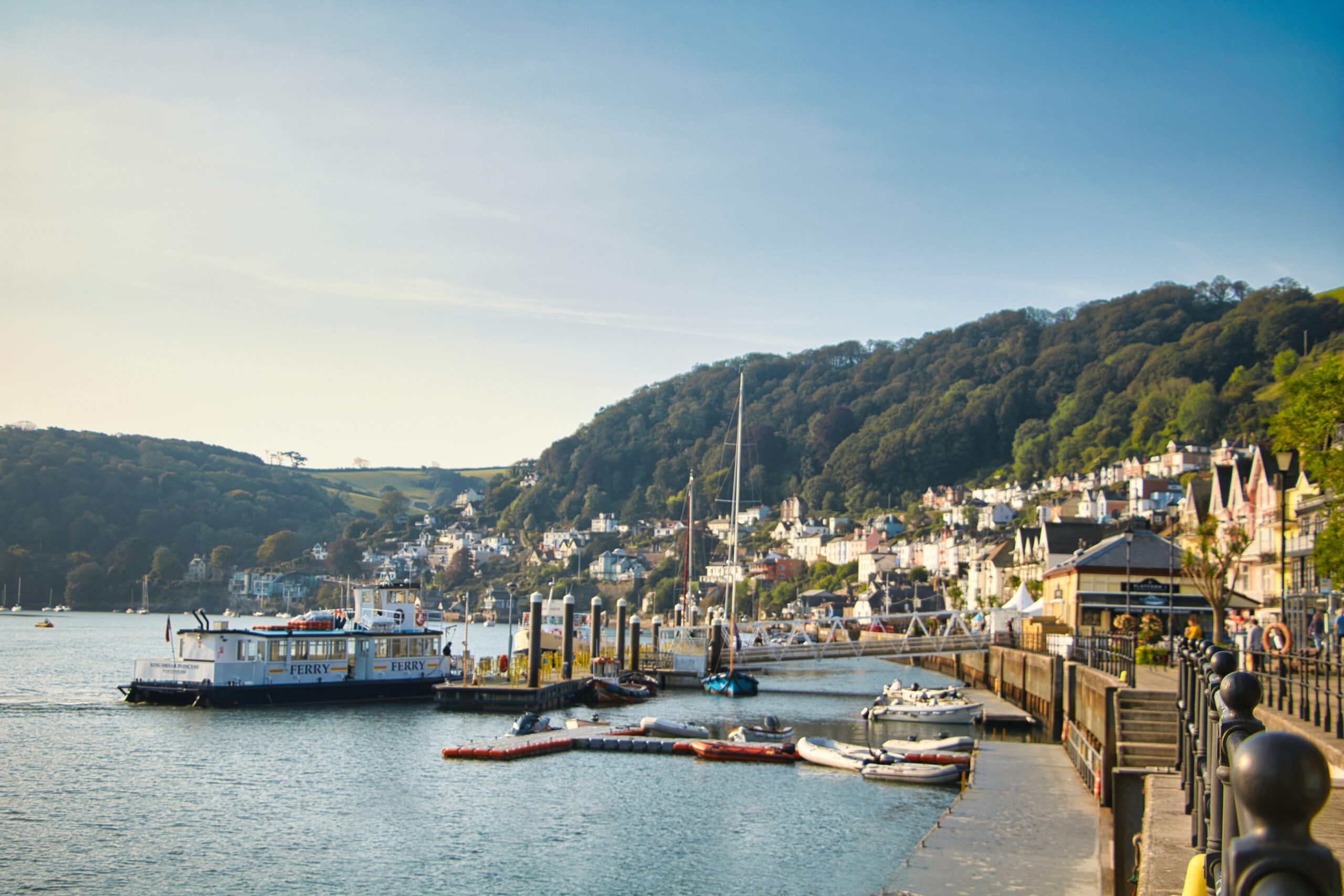 The marina in Dartmouth on a bright sunny evening. Boats docked and the water is still with buildings in the far background.